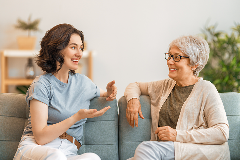 Beautiful mother and daughter are talking and smiling while sitting on couch at home. 